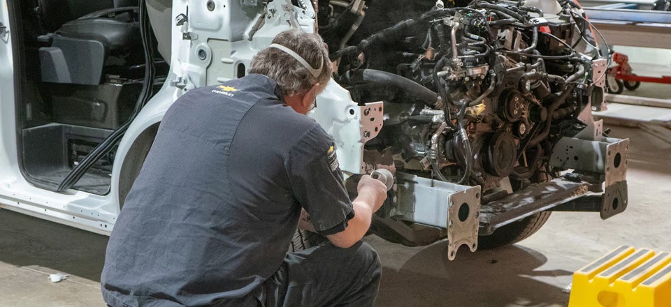 Mechanic in a Chevrolet shirt using a power tool to repair the front frame of a white van with its engine exposed.